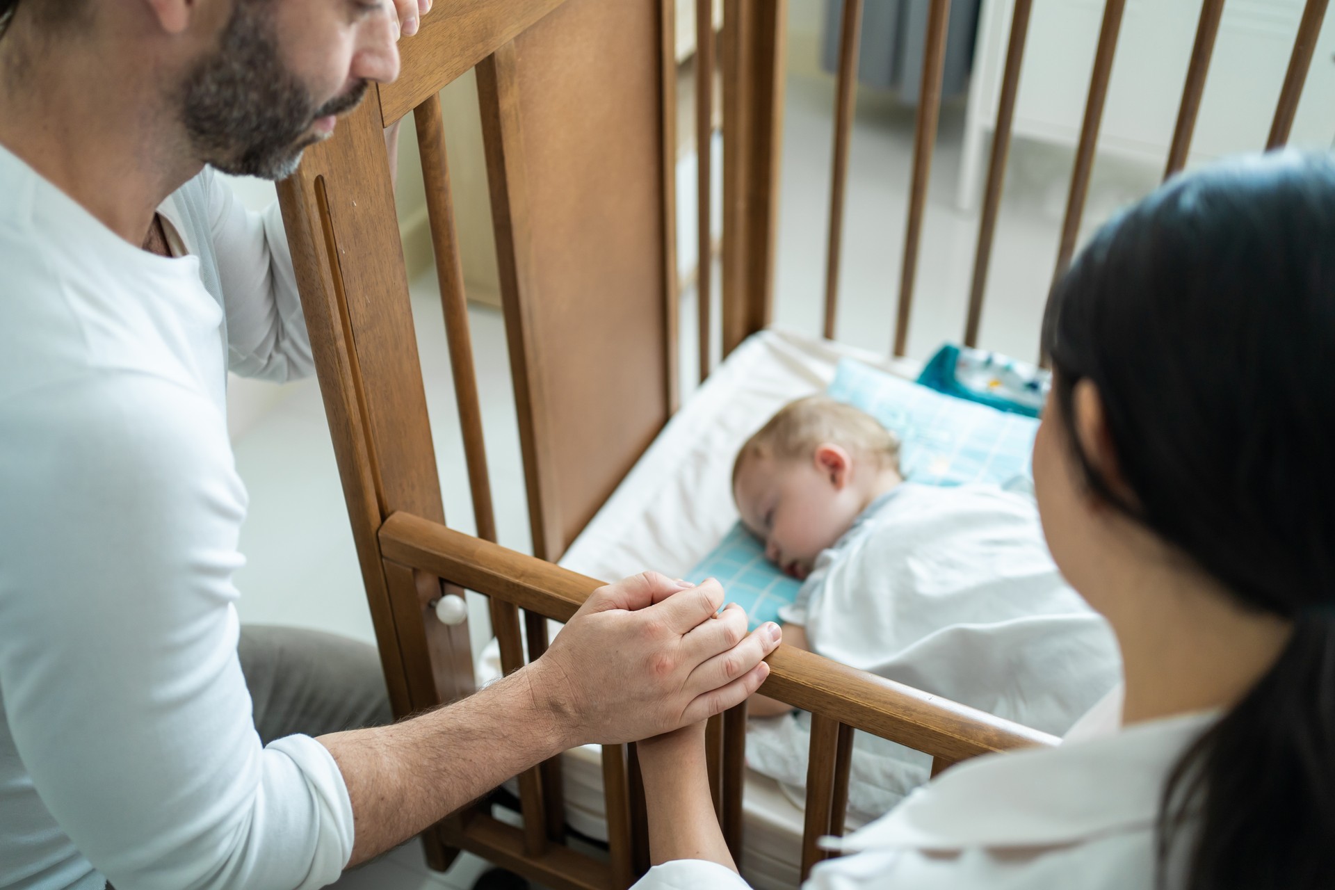 Caucasian parents take care of sleeping baby boy in bedroom at home. Attractive happy family couple, mother and father puts blanket on comforte asleep napping little kid child infant on bed in morning